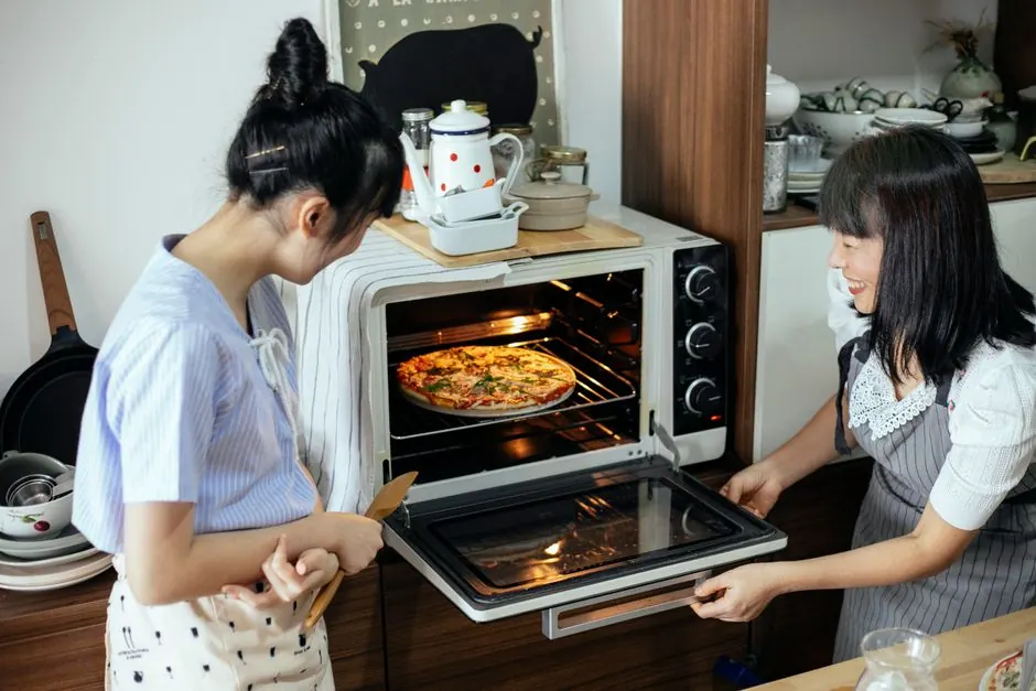 Satisfied Asian ladies in aprons smiling and opening oven while checking yummy homemade pizza made together in modern kitchen